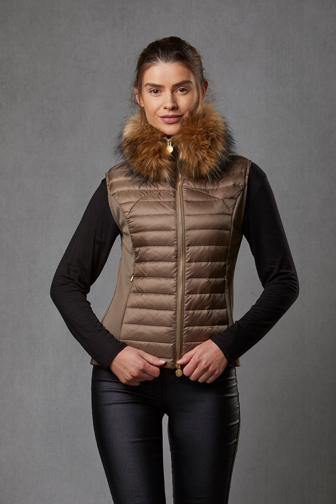 A model is wearing a women's puffer gilet in a khaki, bronze colour. The gilet has a detachable faux fur collar and stretch sides. The puffer gilet has a branded gold zip.