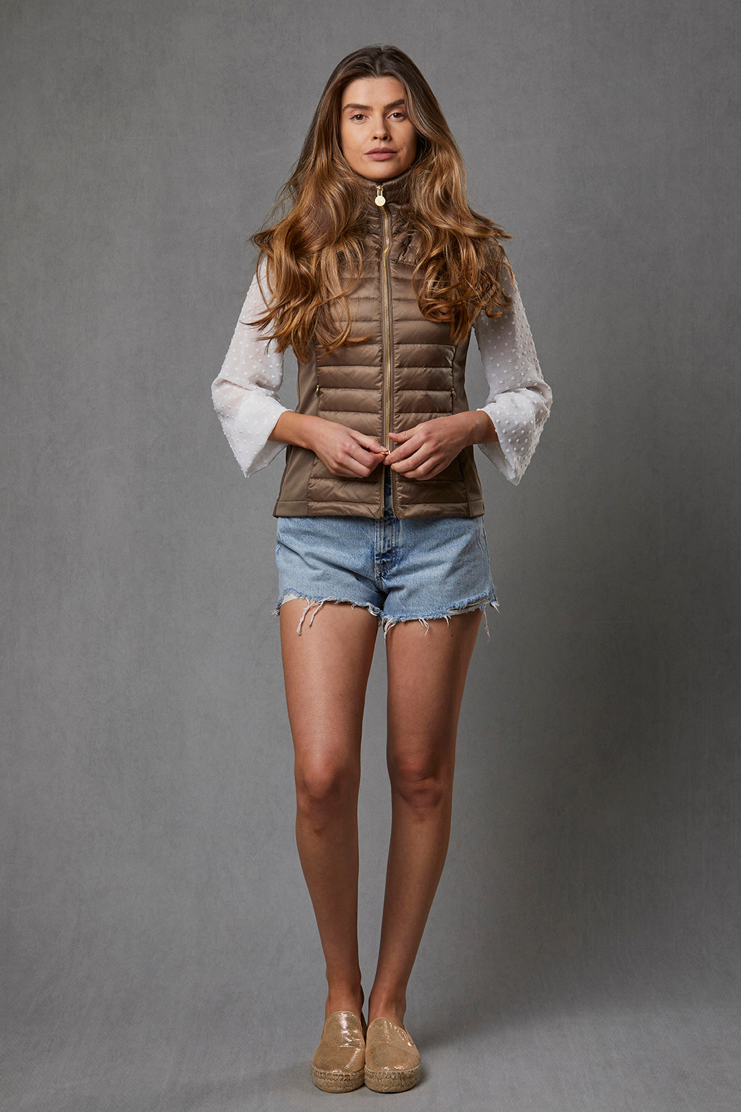Model is wearing a women's puffer gilet in a khaki, bronze colour. The gilet has a detachable faux fur collar which has been removed to show how the puffer gilet can be worn all year round. It has stretch sides and a branded gold zip.
