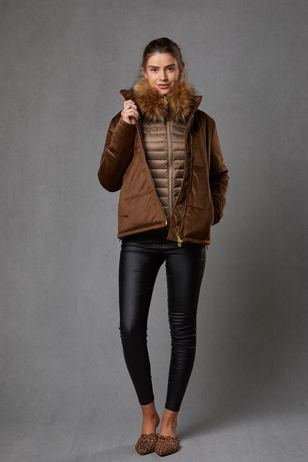 Model wearing a women's puffer gilet in a khaki, bronze colour. The gilet has a detachable faux fur collar and stretch sides. The puffer gilet has a branded gold zip. The model is also wearing a brown puffer jacket over the gilet.