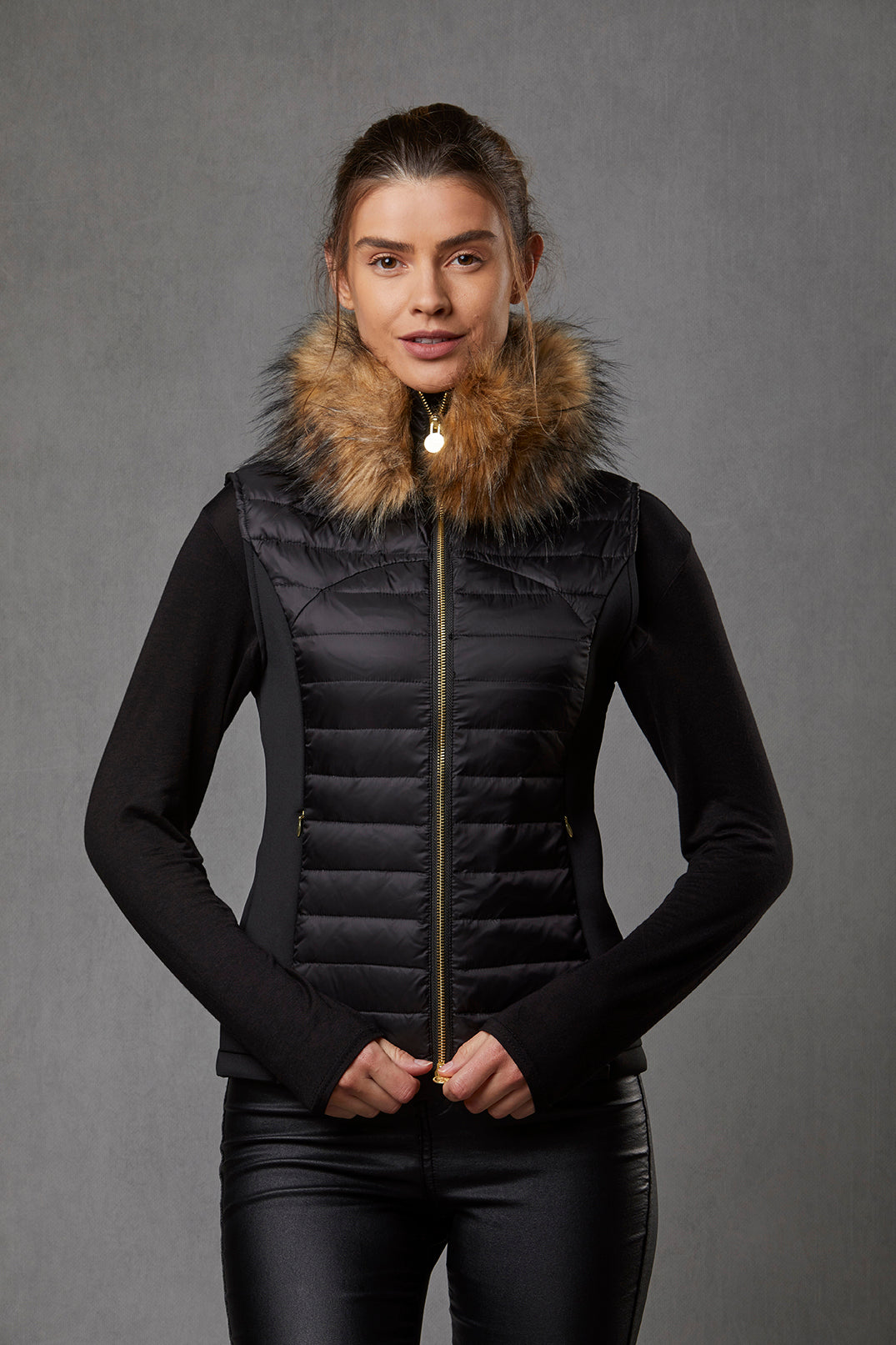 Model wearing a women's puffer gilet in black with stretch sides and a detachable faux fur collar and with gold zip