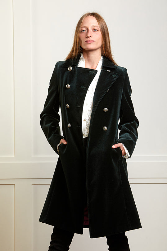 Model wearing a women's mid length coat in dark green velvet. The coat has a classic military style with double breasted silver buttons