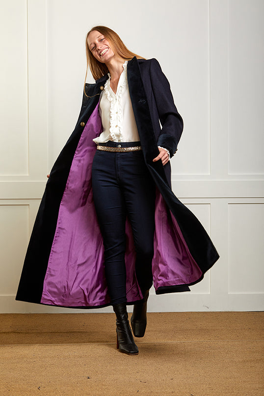Model wearing a women's long blue wool coat in a double breasted style with silver military style buttons. The coat is show open and flowing with a purple lining.