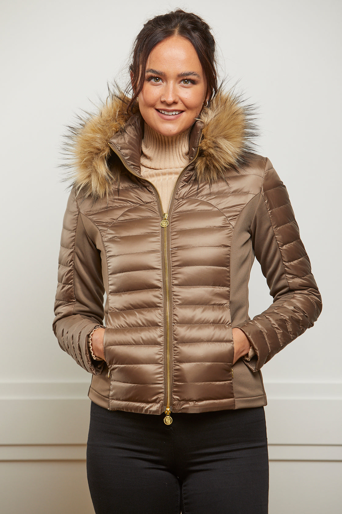 Model wearing a woman's puffer jacket in bronze, khaki colour. It has stretch sides, a gold zip and a detachable faux fur collar.