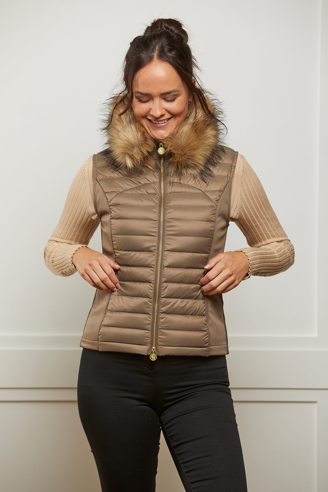 Model wearing a women's puffer gilet in a khaki, bronze colour. The gilet has a detachable faux fur collar and stretch sides. The puffer gilet has a branded gold zip.