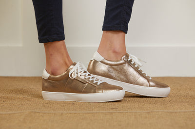 Side view of a model wearing a pair of women's low top leather trainer in bronze gold with white laces and trims