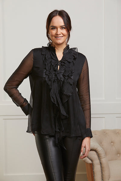 Model wearing a black blouse with a relaxed fit. The blouse has ruffles to the front and sheer pure silk sleeves and a button cuff.